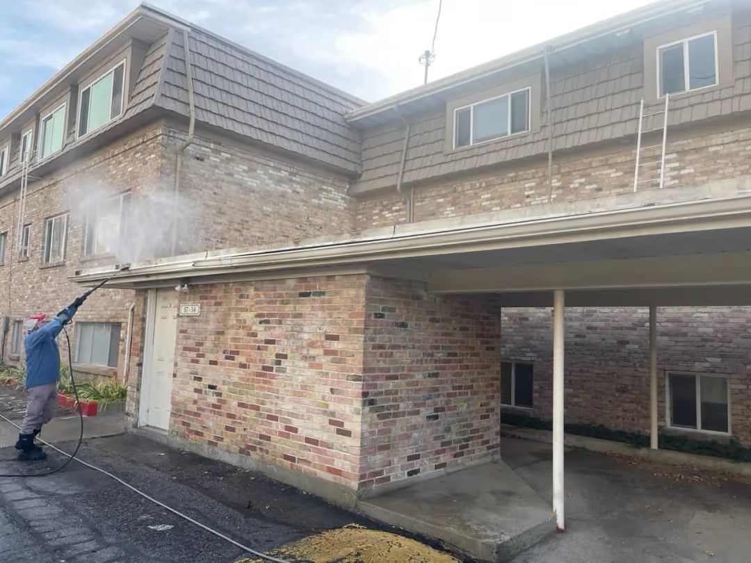 Worker pressure-washing brick house exterior under a covered carport