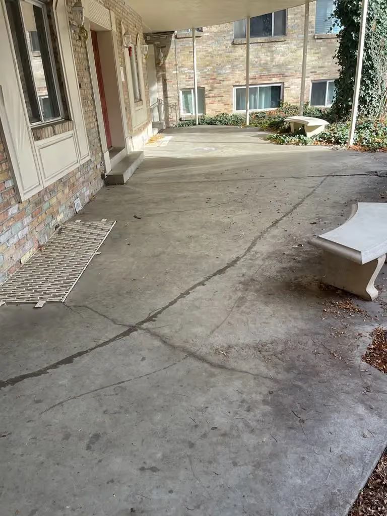Empty covered apartment walkway with concrete floor, brick walls, and a bench on the right.