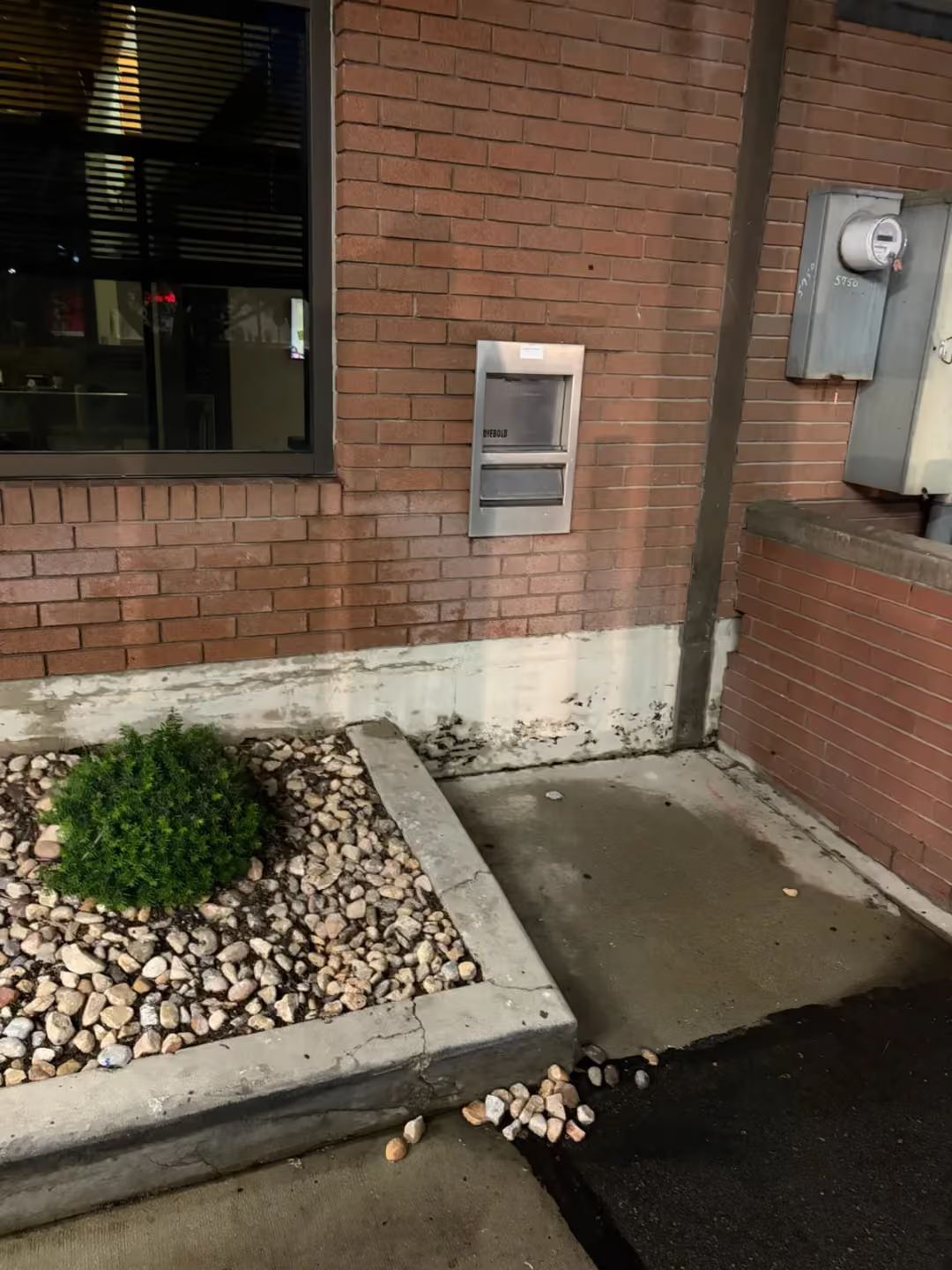 Brick building exterior with a small window well, wall vent, concrete curb, and wet pavement.