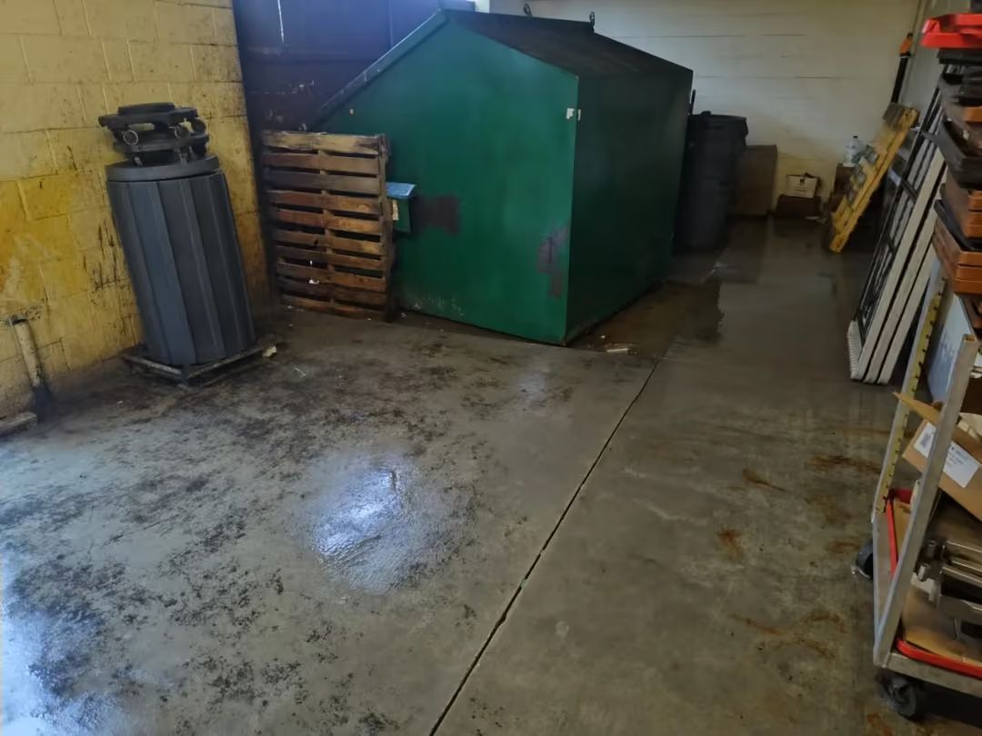 Storage area with a green dumpster, stacked pallets, and shelves in a concrete room.