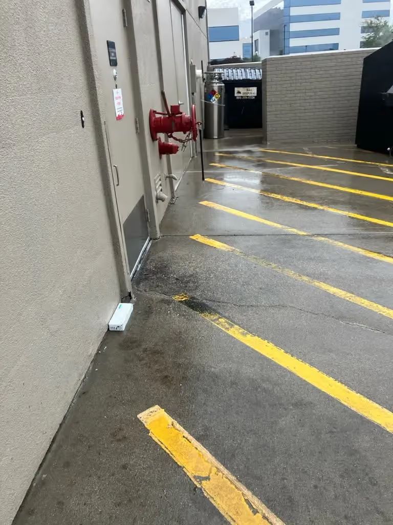 Narrow concrete walkway with yellow diagonal lines, red fire pipes, and building walls in a parking area