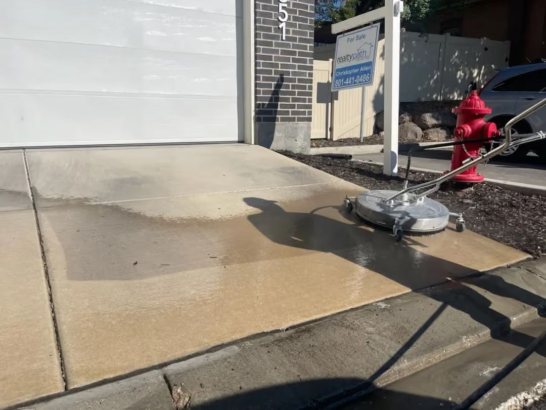Driveway being pressure washed beside a garage, with wet concrete and cleaning equipment visible.