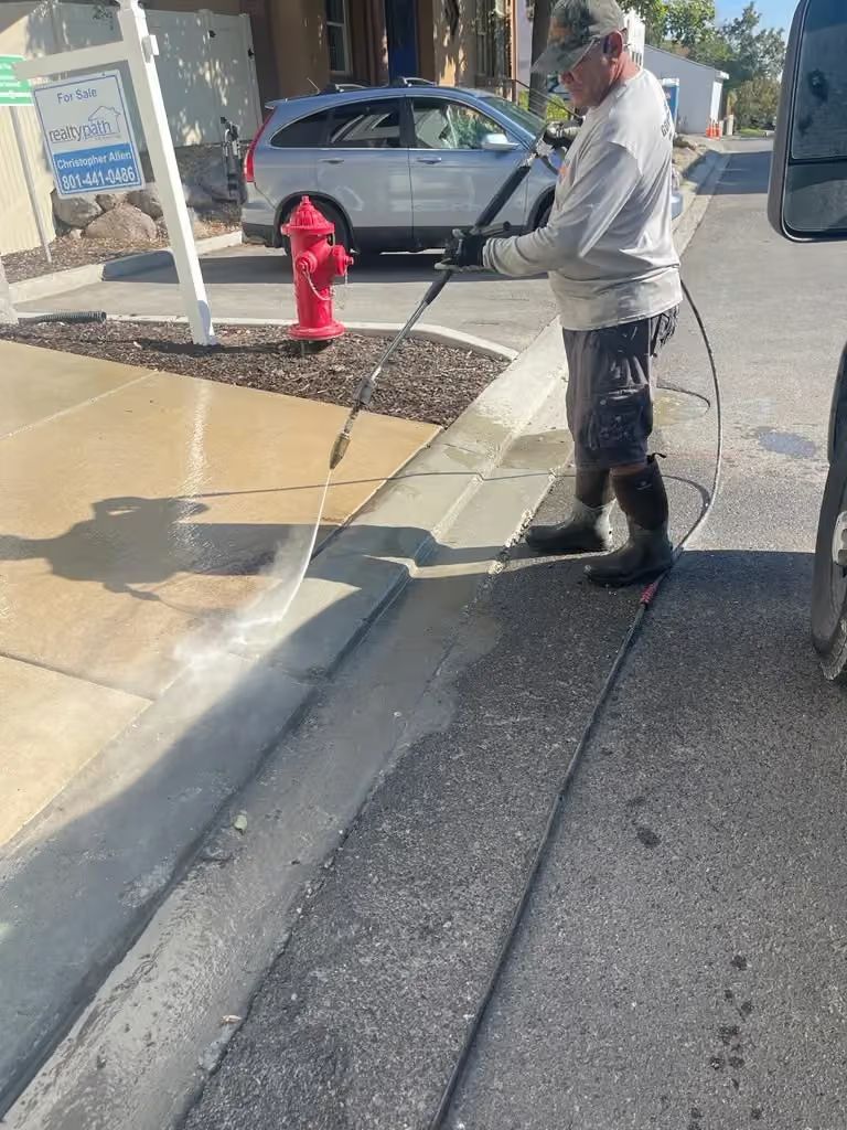 Person washing a sidewalk with a pressure hose beside a red fire hydrant and parked cars