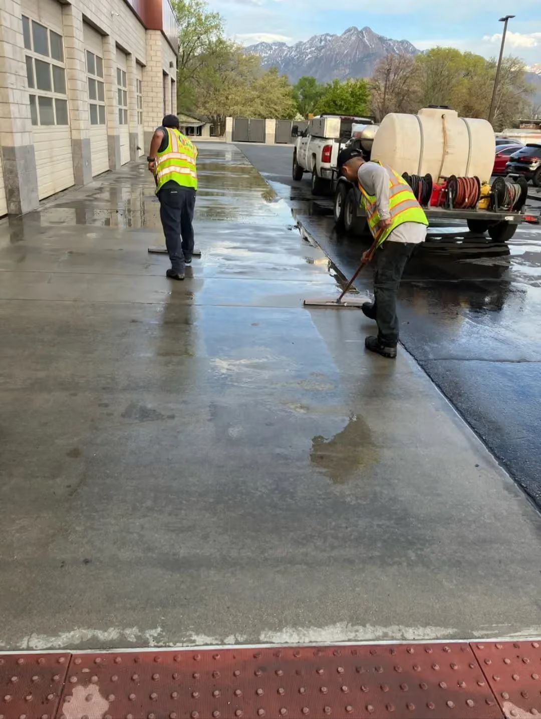 Workers in neon vests cleaning a wet concrete lot beside a building with mountain views.