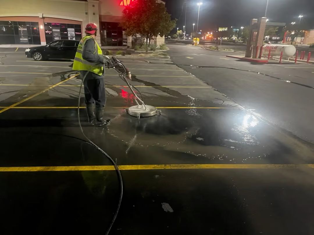 Worker in a reflective jacket stands on a wet street at night beside a hose spraying water across the road.