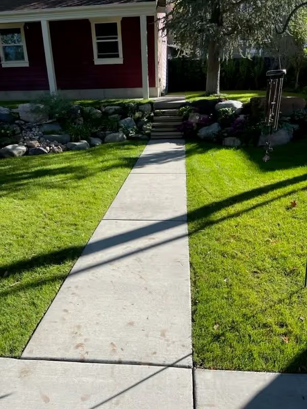 Concrete walkway through a sunlit green lawn leading to a red house with stone landscaping.