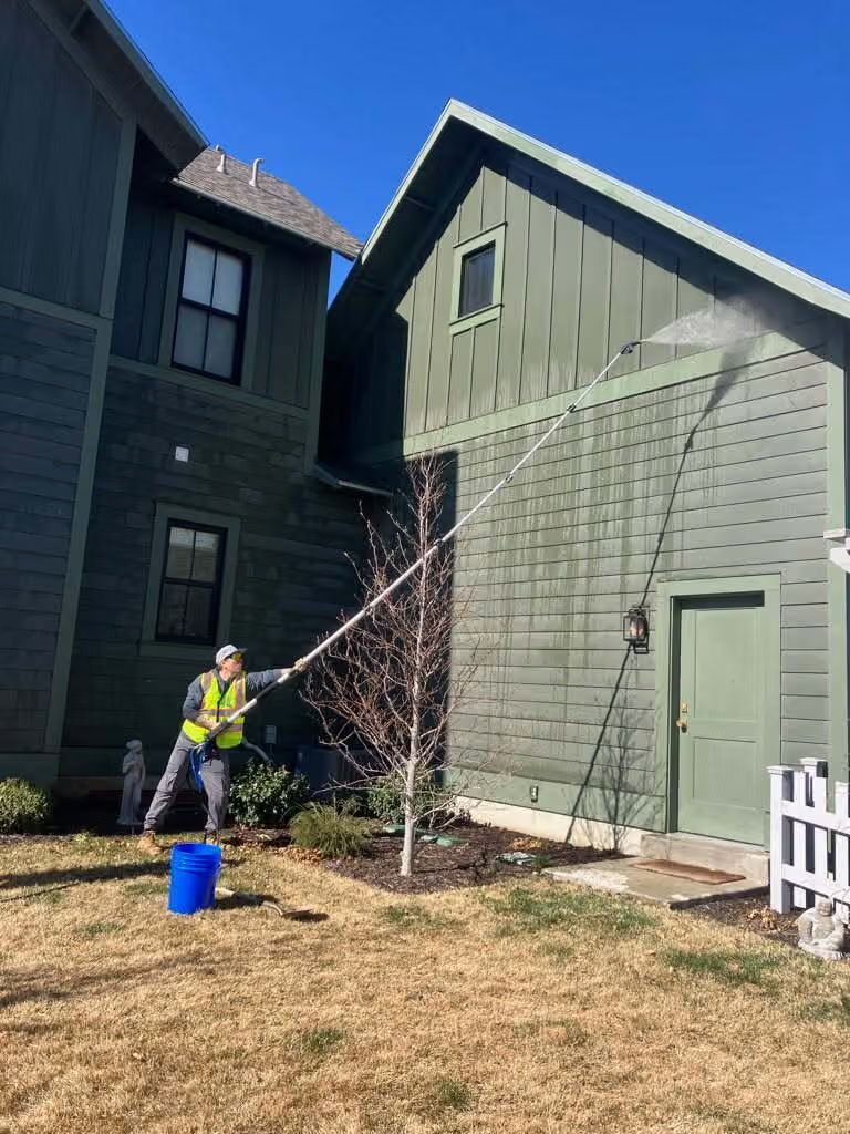 Worker on a ladder power-washing the side of a green house next to a dark house.