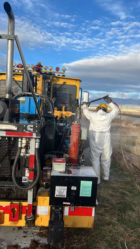 Worker in white protective suit beside industrial machinery on a dusty roadside under a blue sky