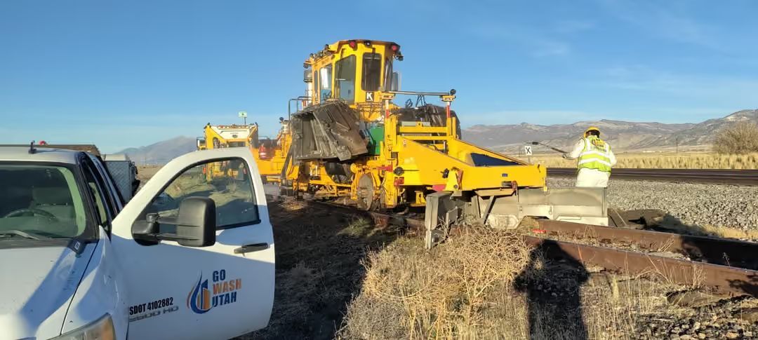 Roadwork scene with yellow construction machinery, a worker, and a white utility truck on a dirt roadside.