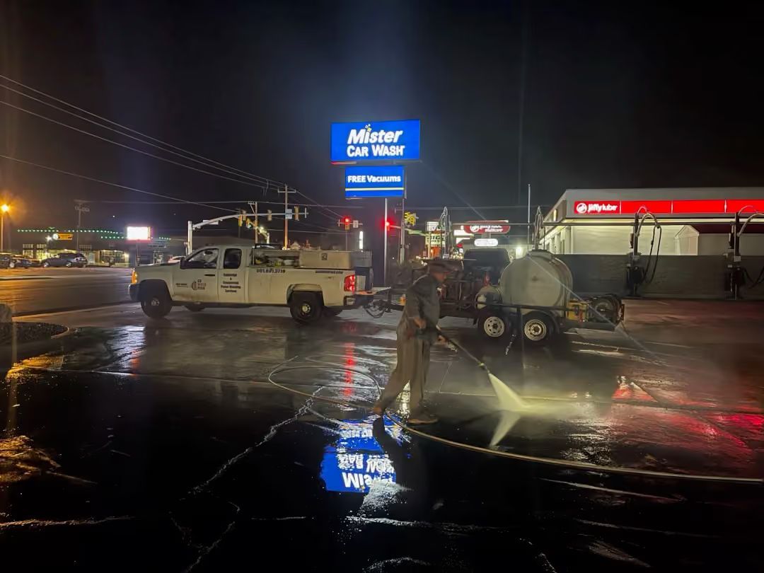 Rainy car lot at night with tow trucks and a bright blue dealership sign reflected in wet pavement