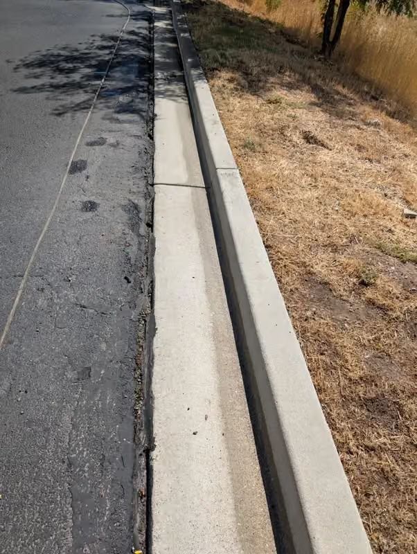 Roadside curb and storm drain along a paved street with dry grass beside it