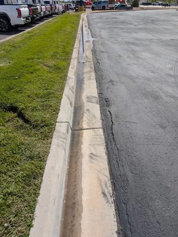 Curved curb and drainage gutter beside a paved parking lot and grassy strip with parked vehicles in the distance