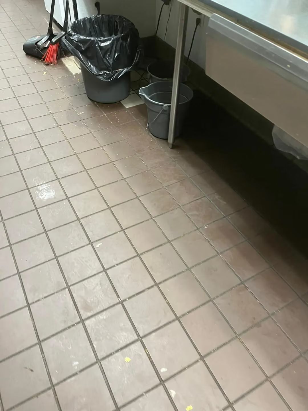 Commercial kitchen floor with tiled floor, trash bags, and cleaning supplies beside a stainless steel counter