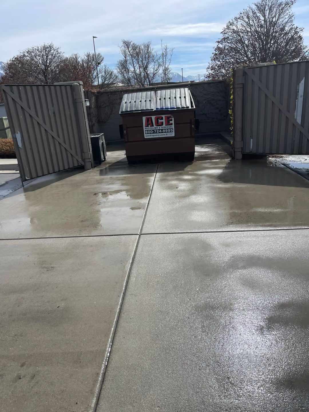 Open dumpster behind a wet concrete lot with metal gates open on both sides