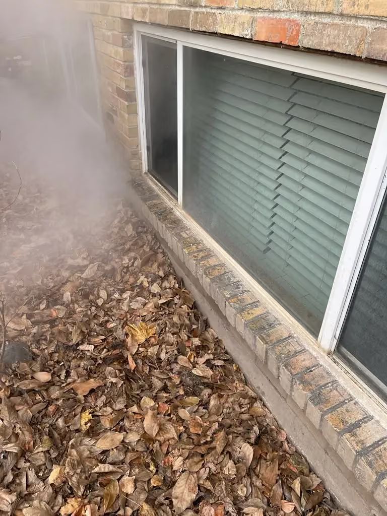 Foggy basement window above a brick foundation, with dry leaves piled along the wall