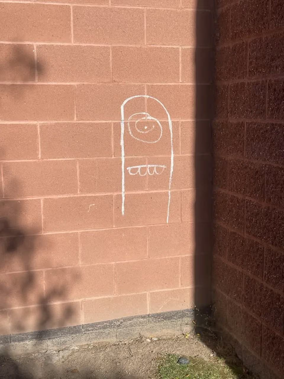 White chalk drawing on a brick wall in sunlight, with a simple cartoon face and torso.