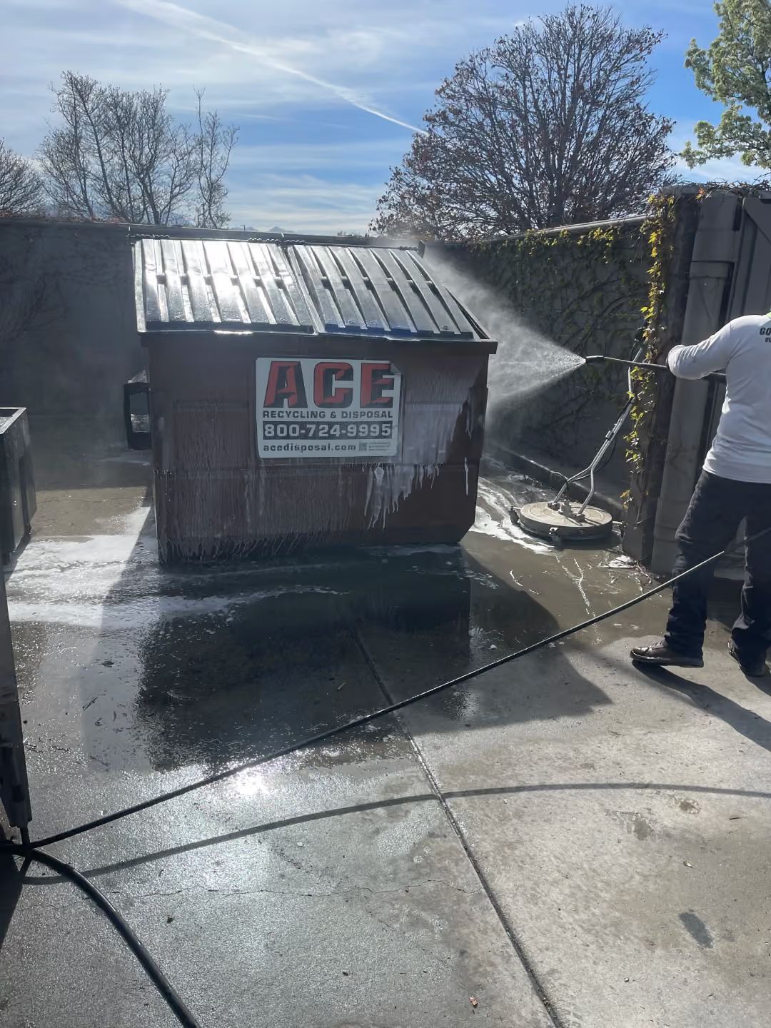 Person pressure-washes an ACE dumpster in a wet outdoor lot.