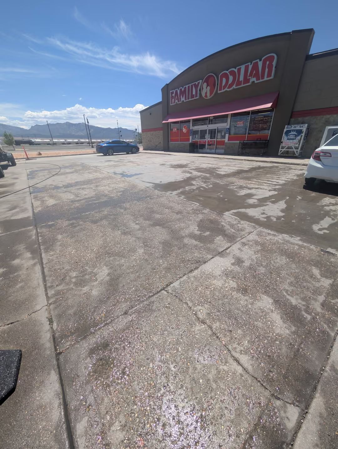 Storefront parking lot on a sunny day, with a large building, red signage, and mountains in the background.