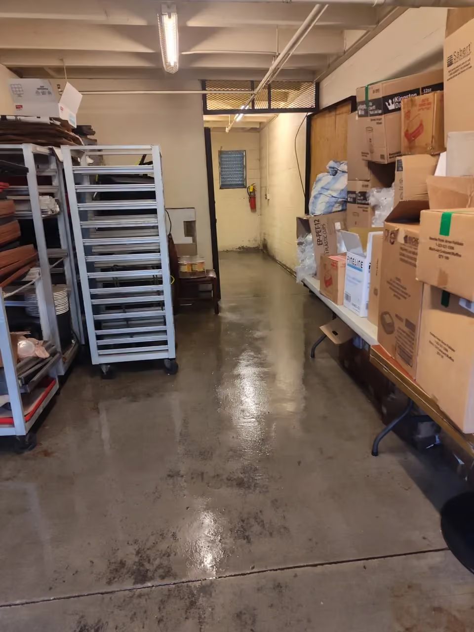 Narrow warehouse aisle with stacked boxes, shelving, and carts on a shiny concrete floor