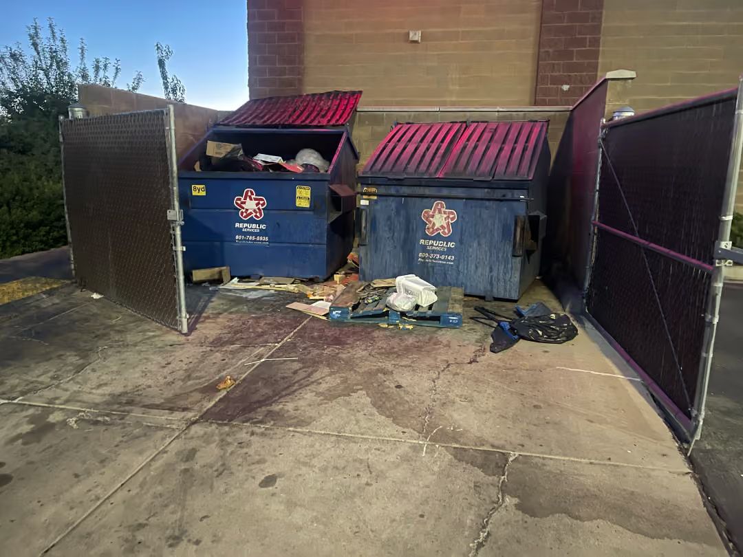 Two overflowing blue dumpsters beside a brick building, with trash scattered on the concrete driveway