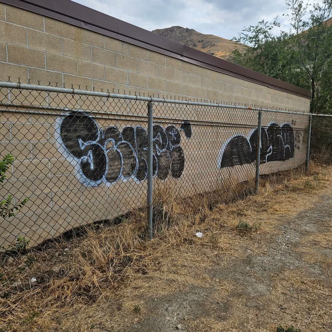 Graffiti on a tan building wall behind a chain-link fence beside a dry path