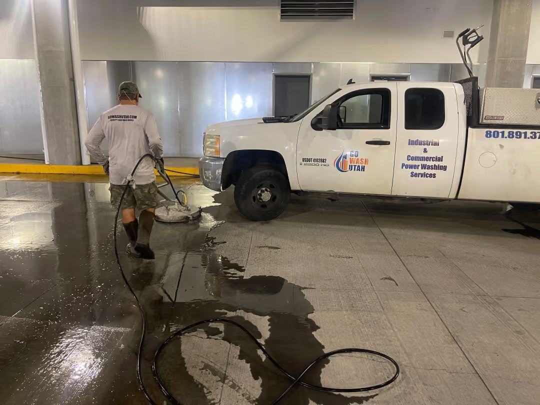 Worker pressure-washing a floor beside a white utility truck in an industrial garage