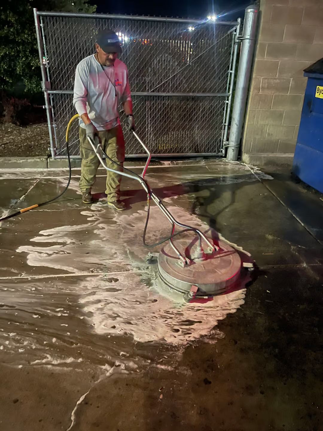 Worker pressure-washing a wet concrete surface beside a chain-link gate at night