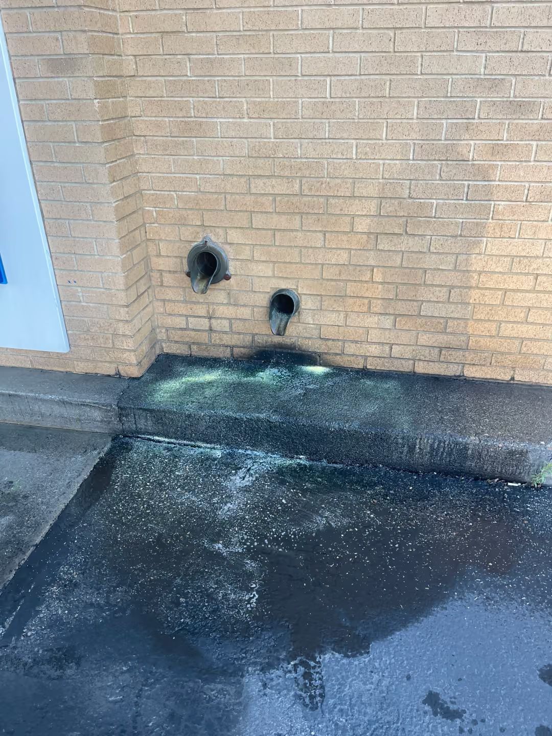 Brick wall with two pipe openings and wet pavement below, showing greenish runoff stain.