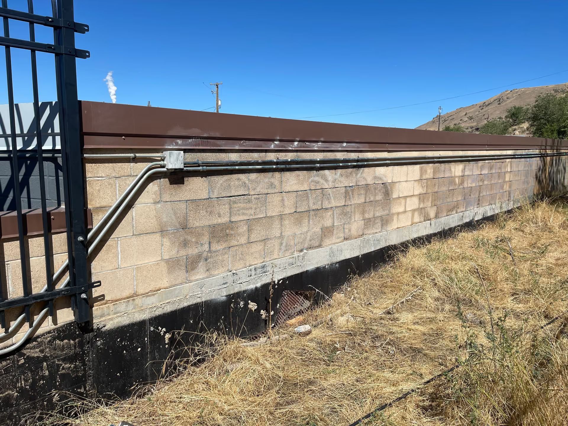 Stone retaining wall beside a black metal gate on a dry hillside under a blue sky