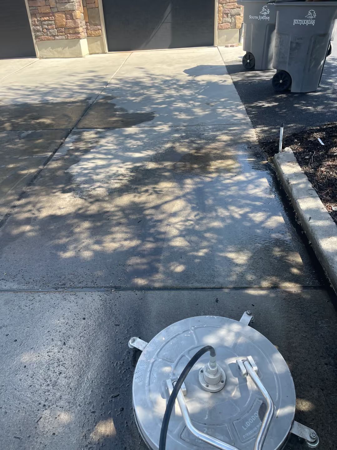 Concrete driveway with a circular utility cover in the foreground and trash bins near a garage.