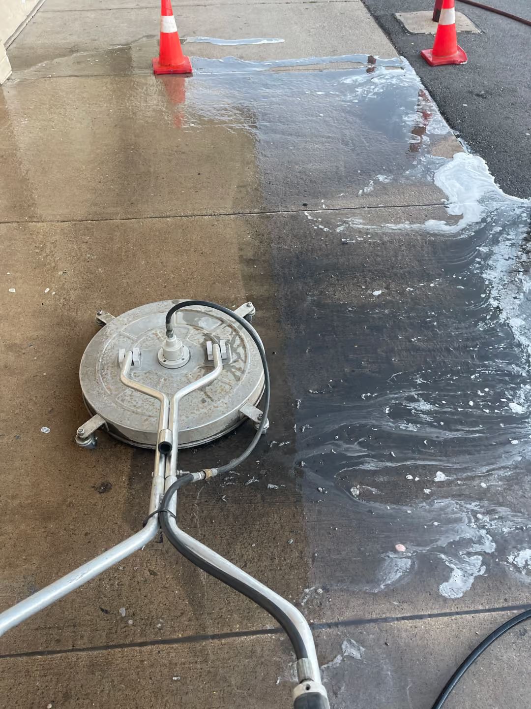 Concrete sidewalk being pressure washed, with wet pavement, foam, and two orange cones.