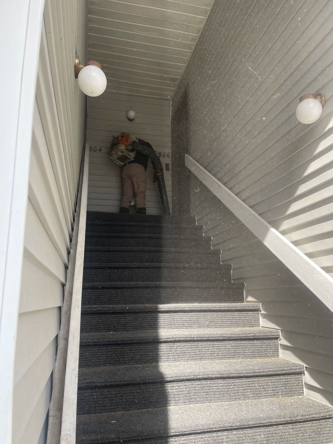 Person carrying items up a narrow outdoor stairwell with white railings and round wall lights