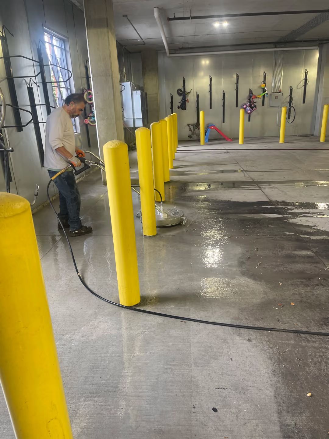 Man cleaning a wet concrete warehouse floor beside bright yellow safety bollards.