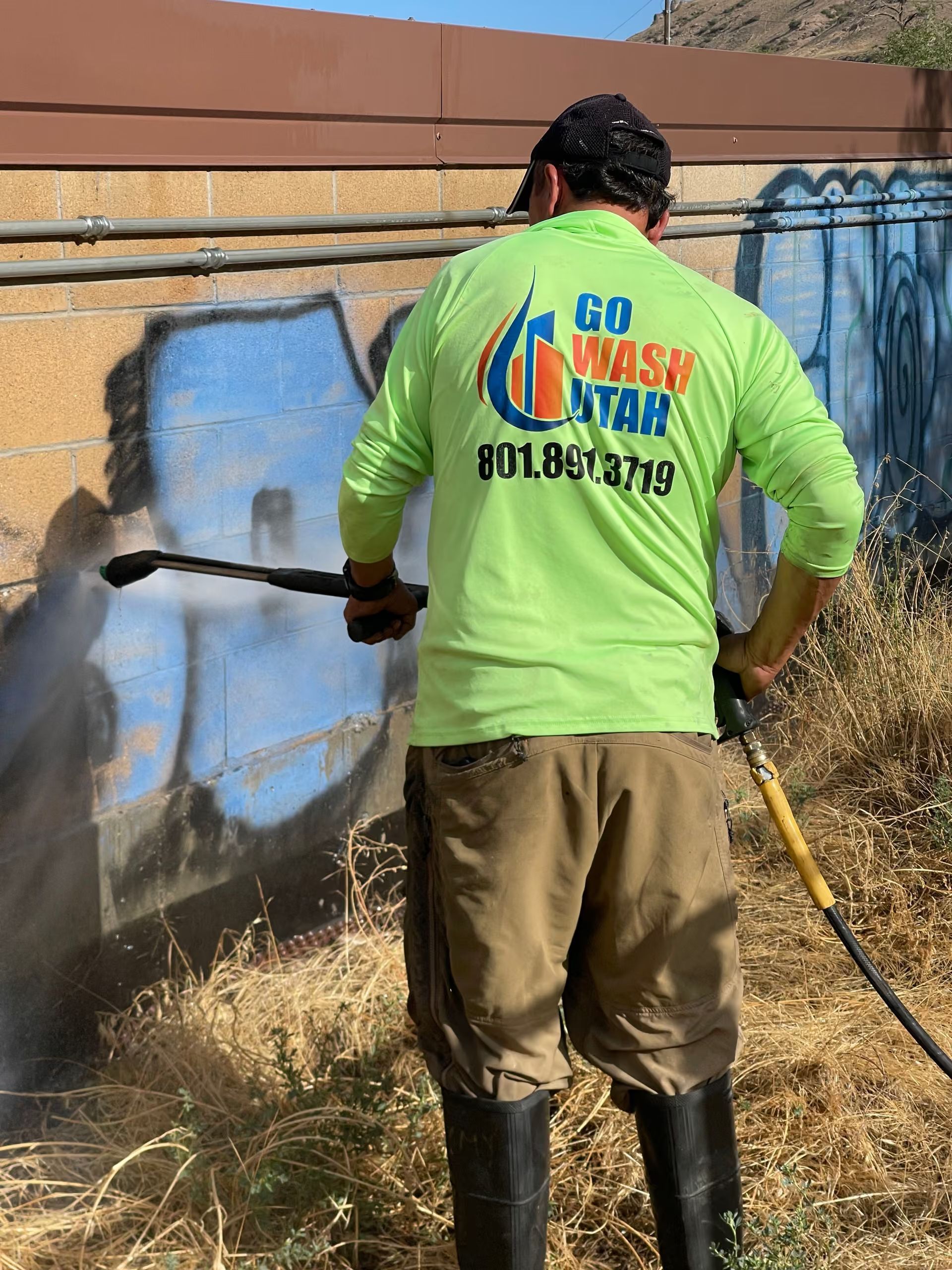 Worker power-washing a blue graffiti-covered wall in a dry outdoor area