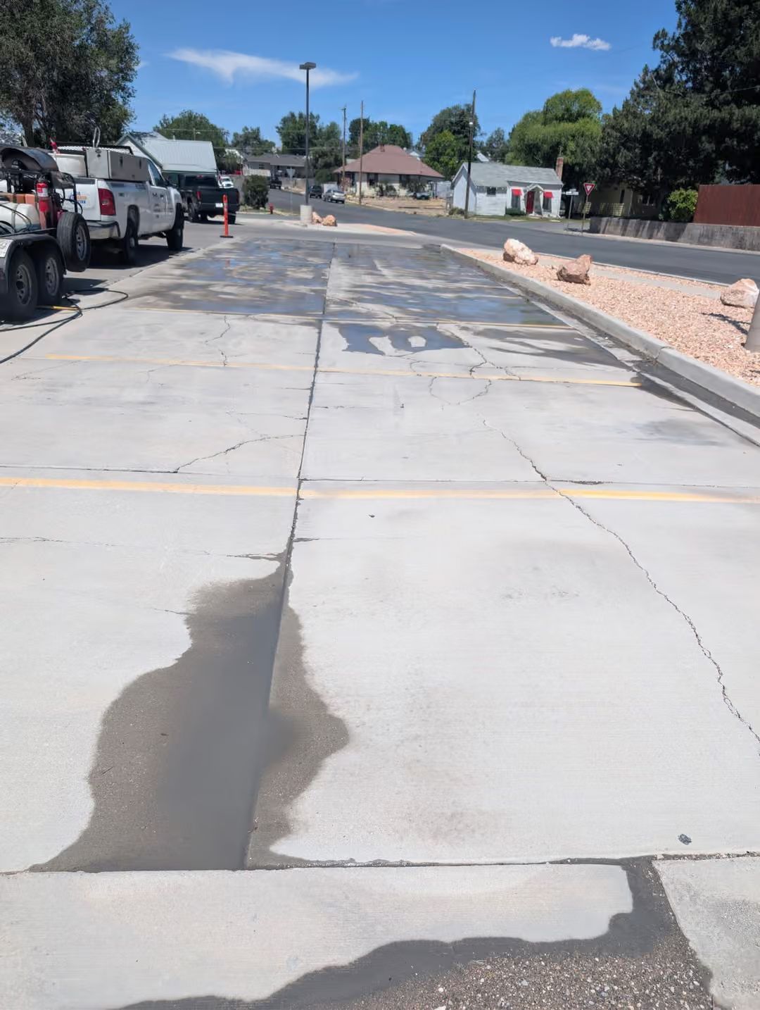 Damaged concrete street under repair, with parked vehicles and orange safety cones along the side.