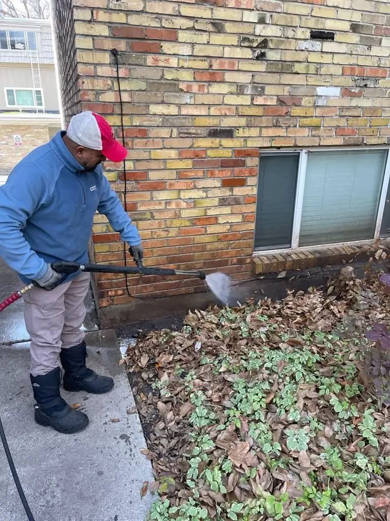 Person using a leaf blower to clear leaves beside a brick building.