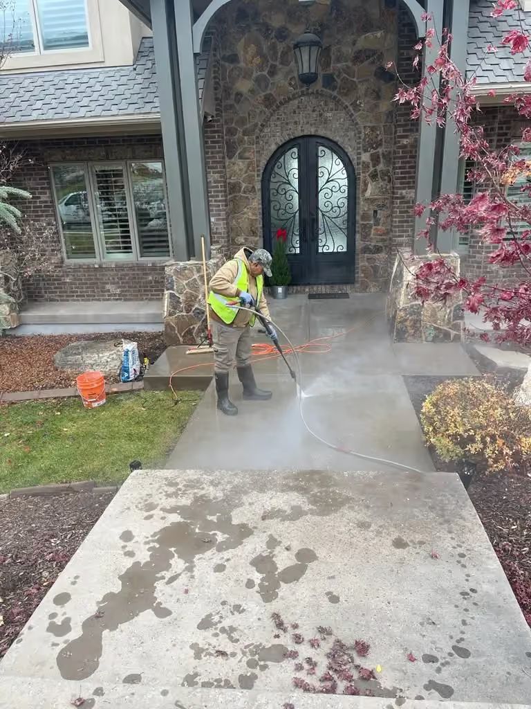 Worker pressure-washing a concrete walkway in front of a stone house with an arched doorway