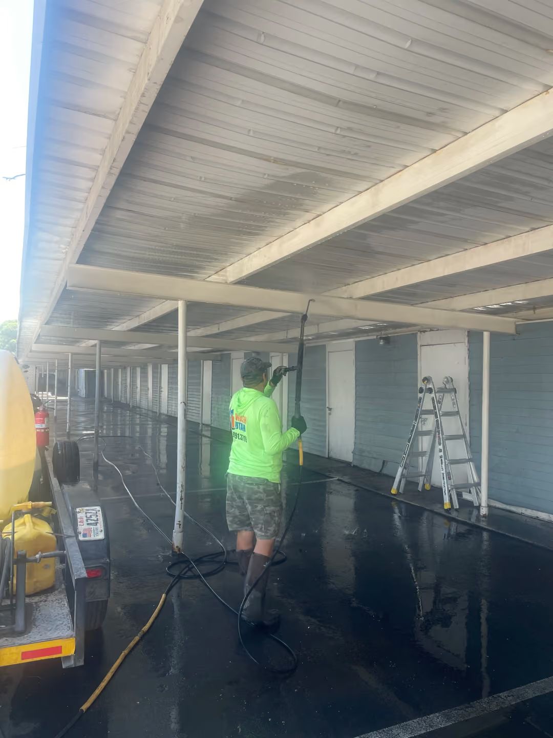 Worker in neon green shirt pressure-washing a covered walkway with wet concrete floor and ladders nearby