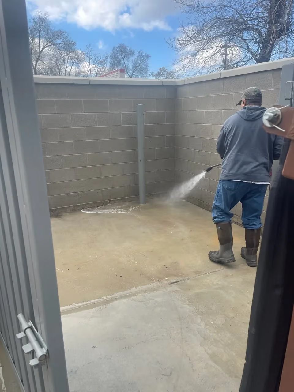Person pressure washing a concrete courtyard beside gray block walls on a sunny day
