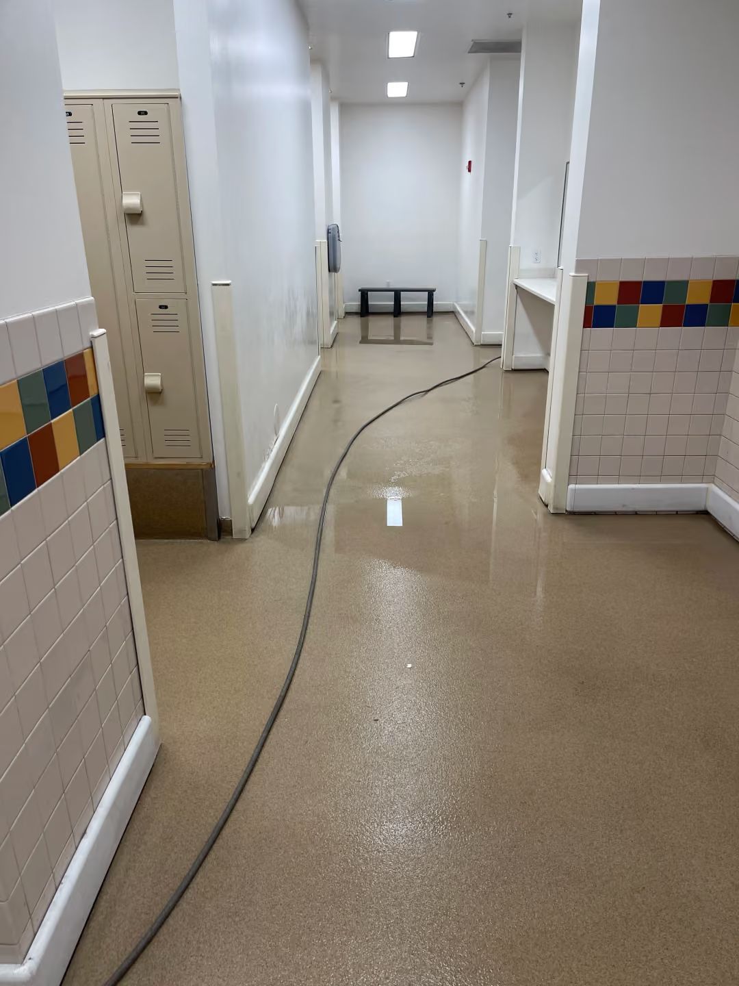 Long hallway with beige floor, white walls, lockers, and a black cable lying across the walkway.