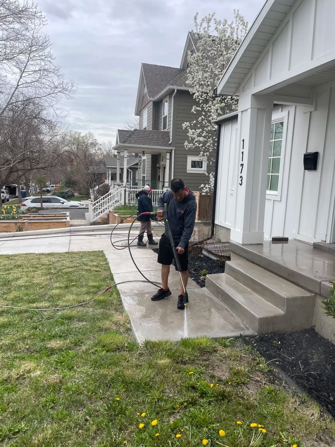 Two people using long-handled tools on a sidewalk beside houses and a grassy lawn