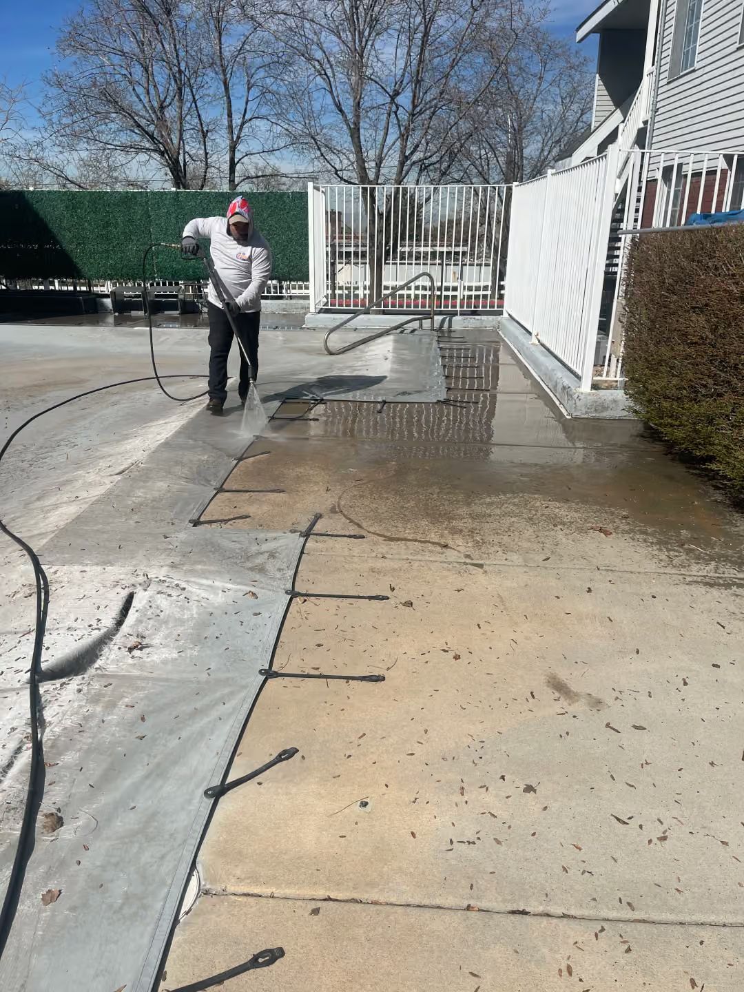Person pressure-washing a concrete driveway beside a house and fence under a clear sky