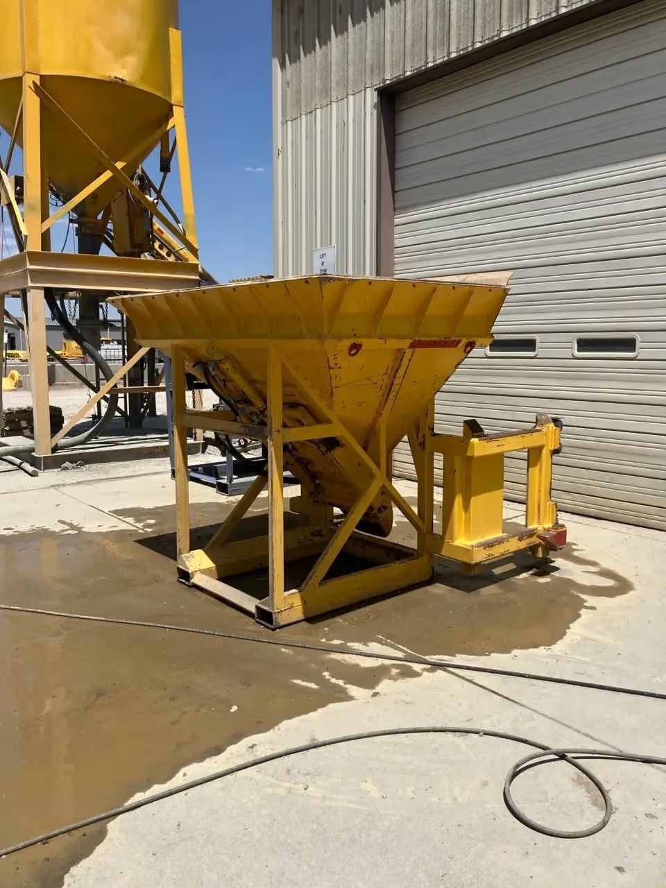 Yellow industrial hopper and conveyor equipment beside a warehouse roll-up door on a concrete lot.