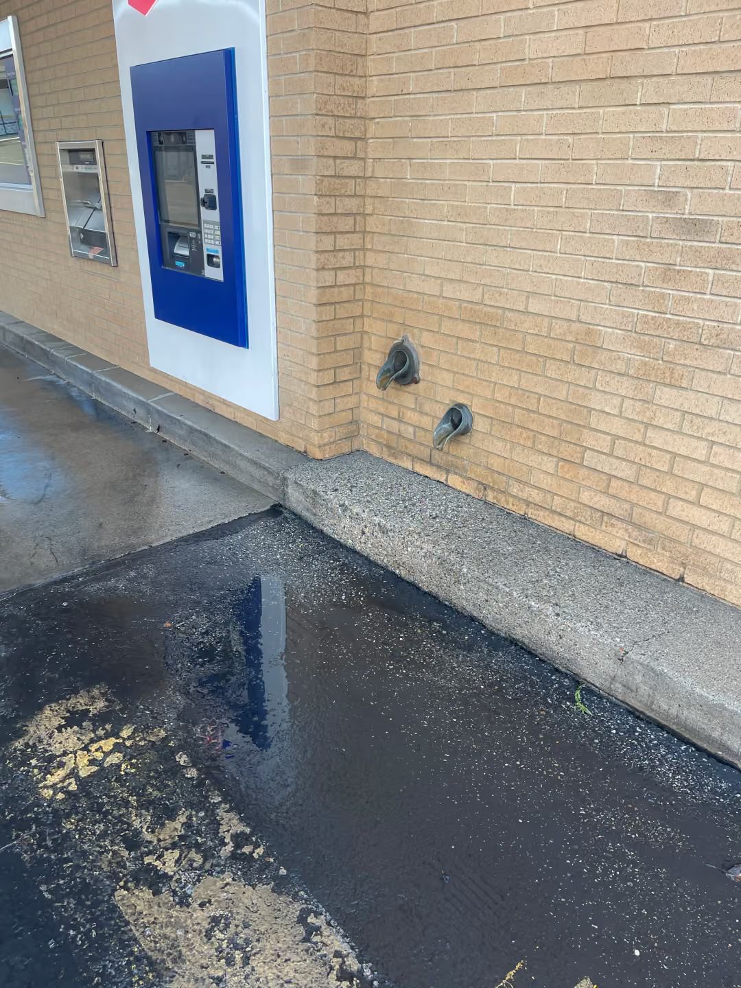Drive-thru ATM in a blue frame on a brick wall beside a wet pavement and puddle.
