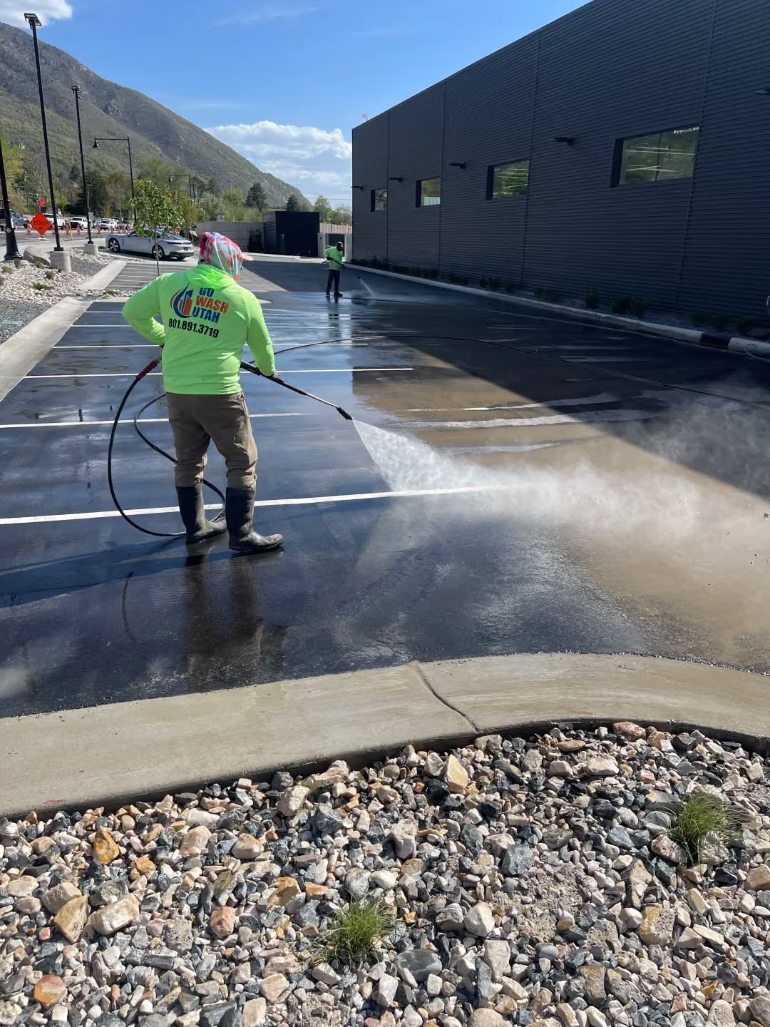 Worker spraying water on a paved surface beside a dark building on a sunny day