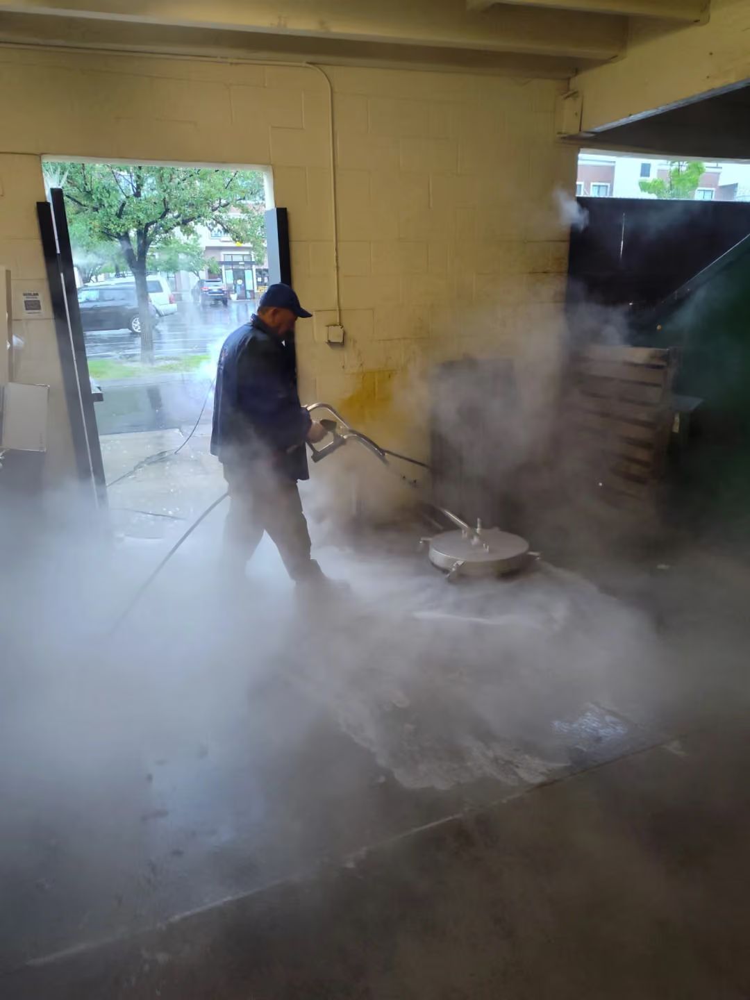 Worker pressure-washing a smoky floor inside a garage, with bright daylight coming through the open door.
