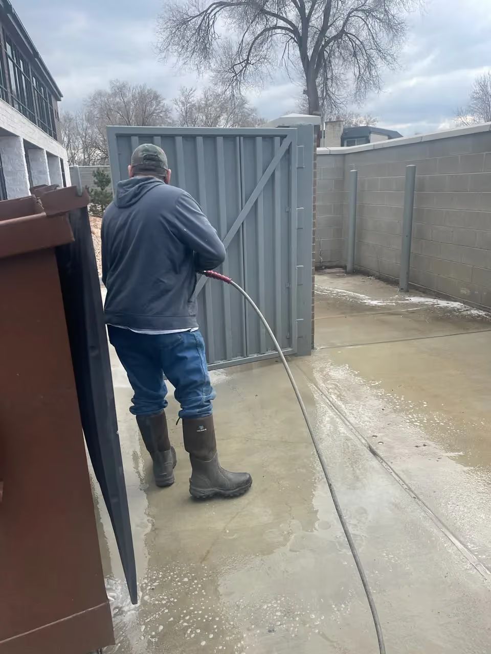 Person pressure washing a concrete patio near a gray fence on a cloudy day