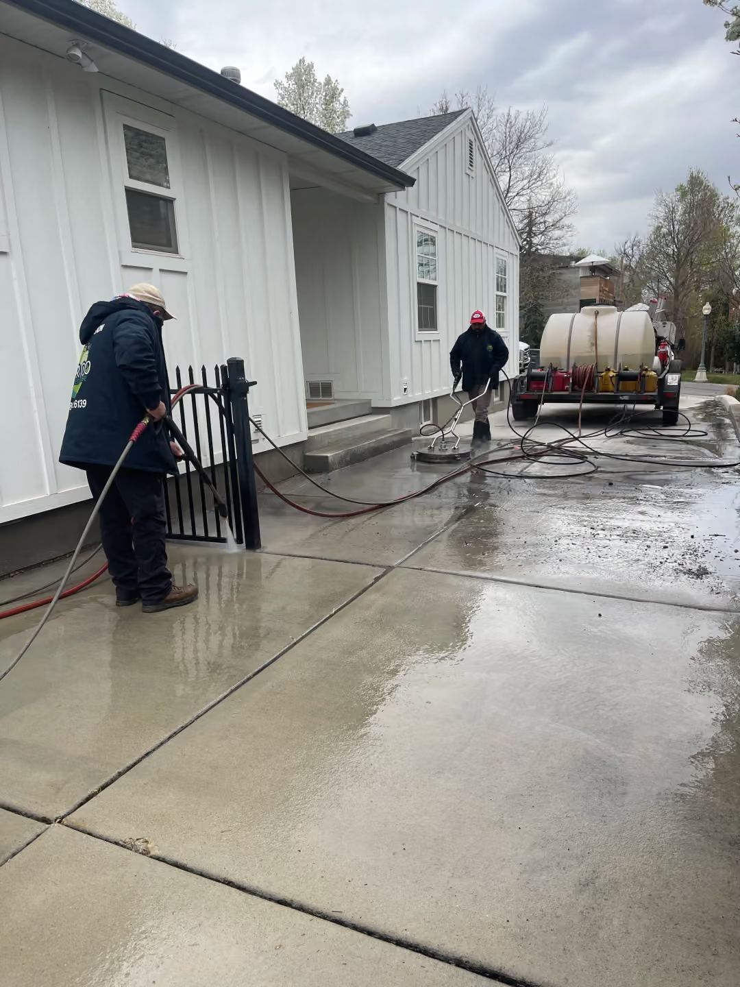 Workers spraying concrete sealant beside a white house on a wet driveway