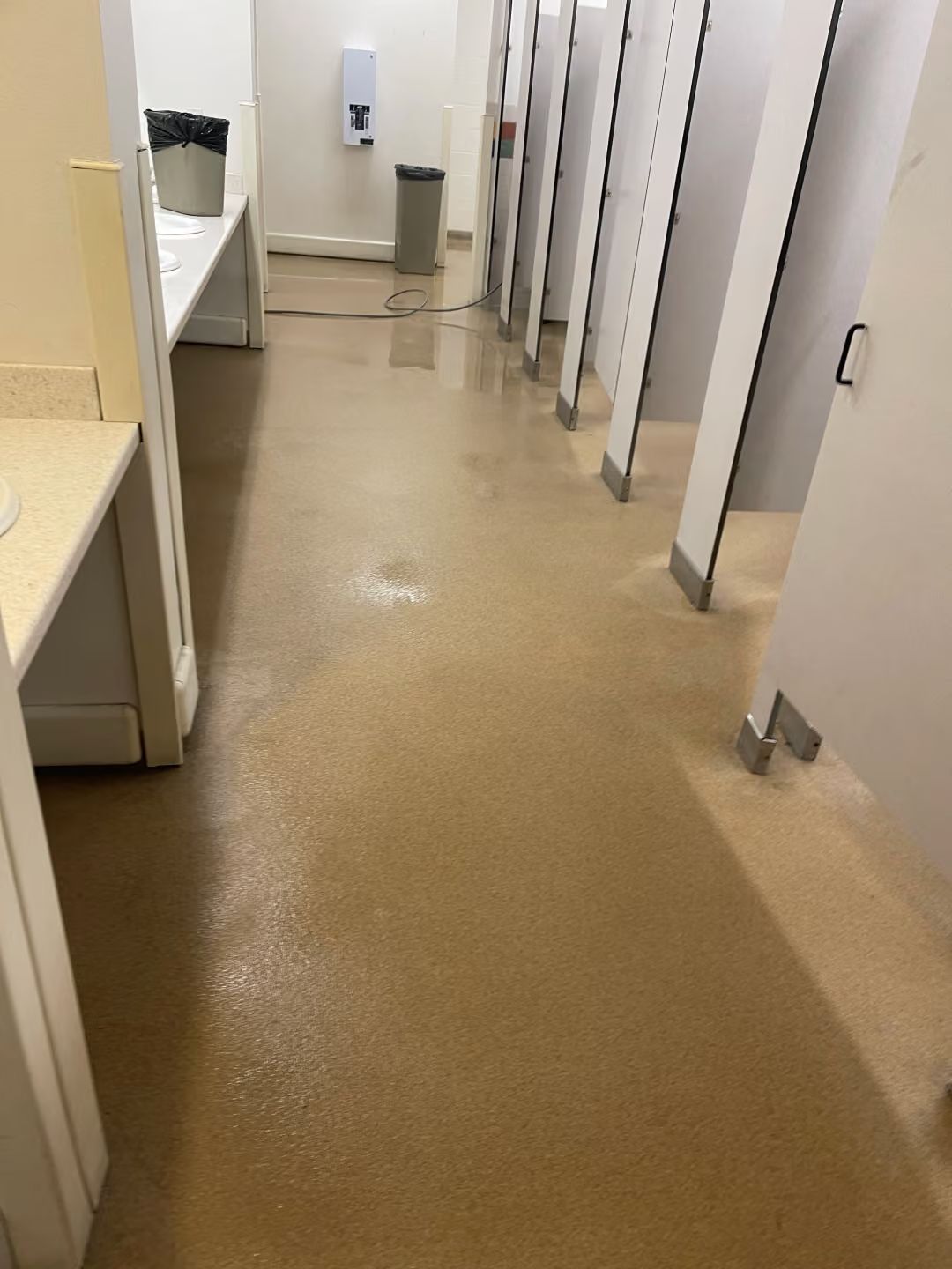 Narrow restroom corridor with beige floor and white stall partitions, wet surface reflecting light