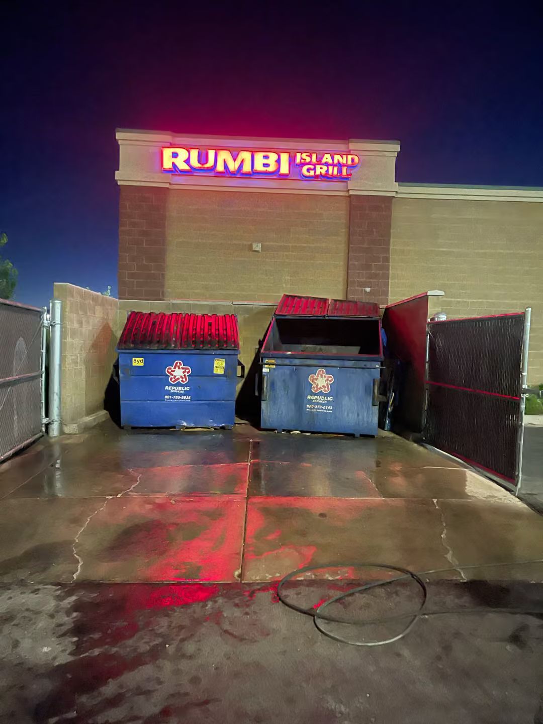 Two blue dumpsters with red lids in a wet alley outside a brick building with a neon sign.